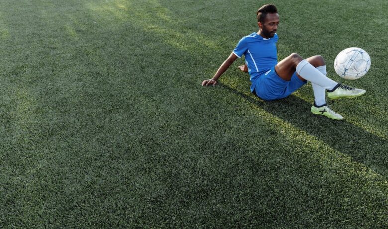 Homem sentado em campo de grama sintética, em traje de futebol azul, controlando uma bola.